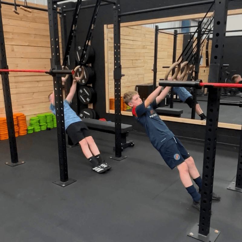 Two boys perform inverted rows on a squat rack in a gym, pulling their bodies toward the bar while feet hover off the floor near weight plates.