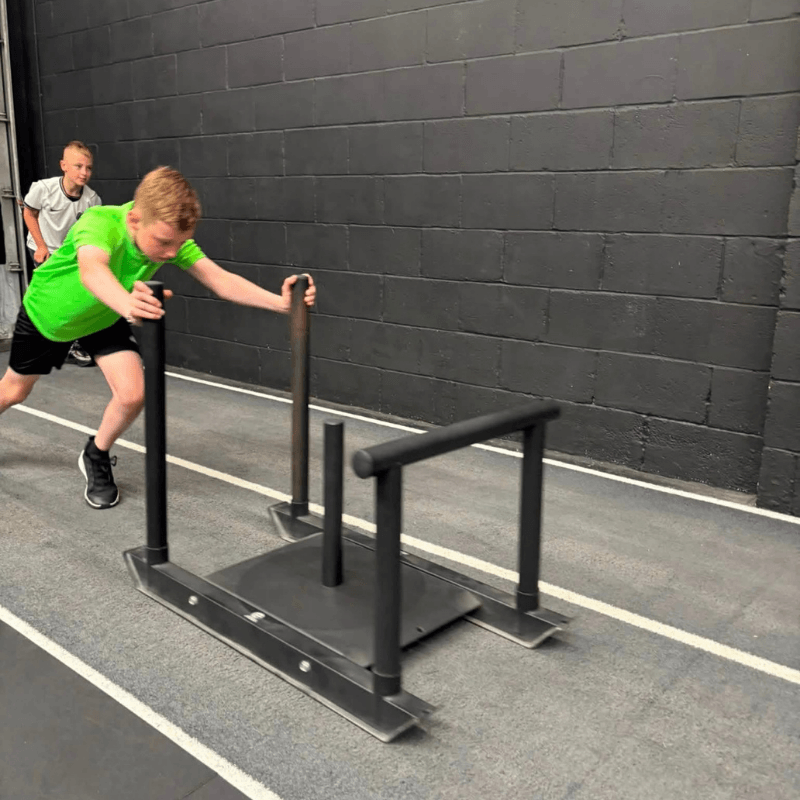 Boy in a bright green shirt pushes a black sled with handles across a gym floor, with another boy watching in the background on a dark brick wall.